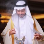 A Middle Eastern man in traditional white and gold attire holding an ornate incense burner indoors.