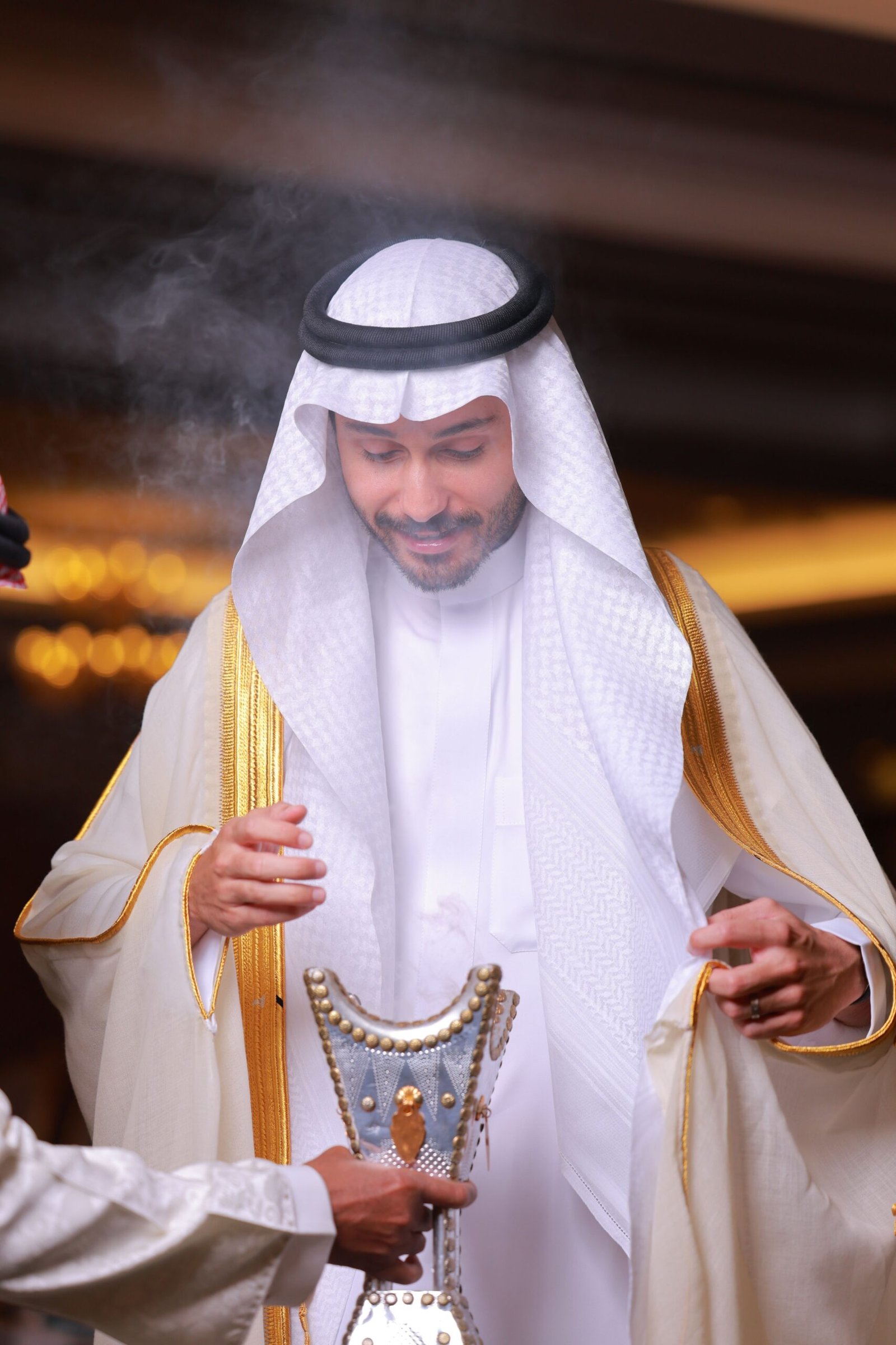 A Middle Eastern man in traditional white and gold attire holding an ornate incense burner indoors.
